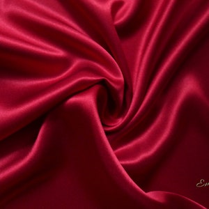 May include: A close-up of a luxurious, deep red satin fabric with soft folds and a swirling pattern. The fabric has a subtle sheen, reflecting light and creating highlights and shadows. The word "Emsilk" is in the bottom right corner.