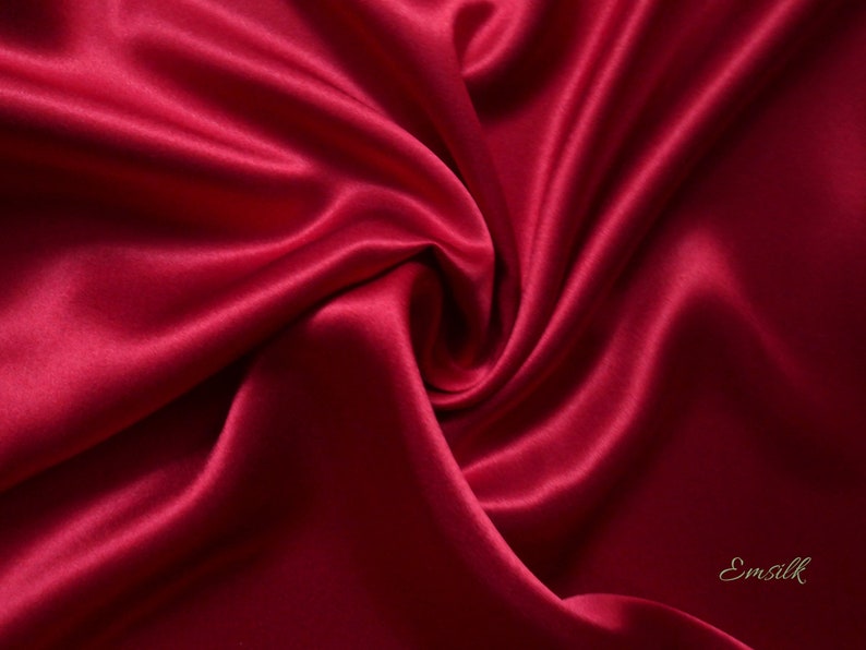 May include: A close-up of a luxurious, deep red satin fabric with soft folds and a swirling pattern. The fabric has a subtle sheen, reflecting light and creating highlights and shadows. The word "Emsilk" is in the bottom right corner.