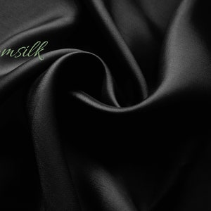 May include: Black satin fabric with a smooth, silky texture. The fabric is draped and folded, creating soft curves and folds. The word "Emsilk" is written in green text on the fabric.