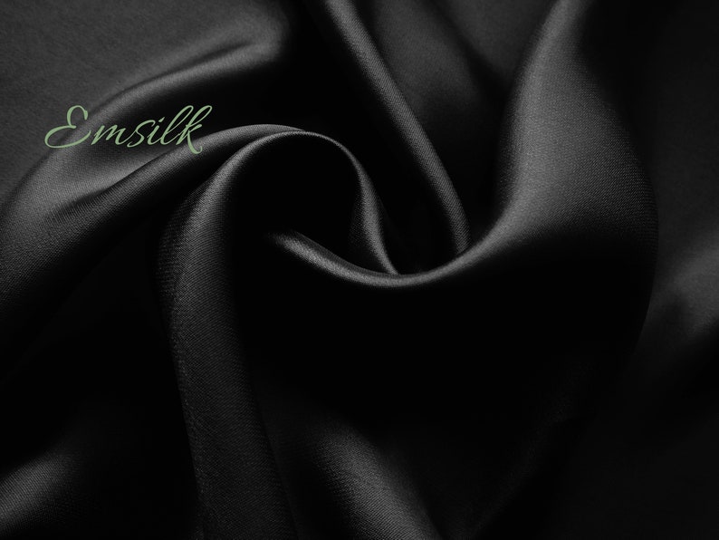 May include: Black satin fabric with a smooth, silky texture. The fabric is draped and folded, creating soft curves and folds. The word "Emsilk" is written in green text on the fabric.