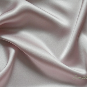 May include: A close-up of a smooth, pale pink silk fabric with a subtle sheen. The fabric is draped and folded, creating soft curves and folds. The text "Emsilk" is visible in the lower right corner.