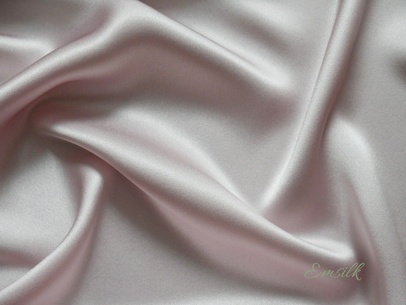 May include: A close-up of a smooth, pale pink silk fabric with a subtle sheen. The fabric is draped and folded, creating soft curves and folds. The text "Emsilk" is visible in the lower right corner.