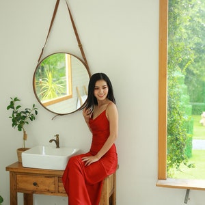 May include: A woman wearing a red dress sits on a wooden vanity with a white porcelain sink. She is looking at the camera and smiling. There is a round mirror with a leather strap hanging on the wall behind her.