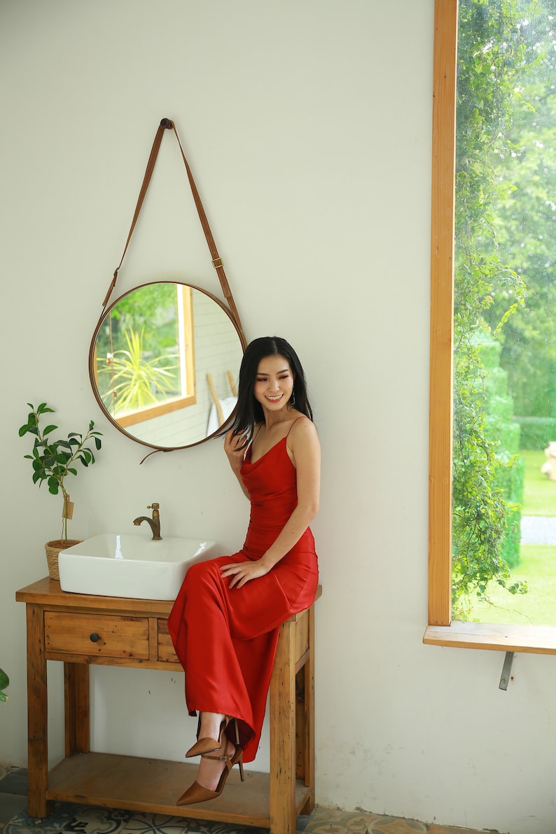 May include: A woman wearing a red dress sits on a wooden vanity with a white porcelain sink. She is looking at the camera and smiling. There is a round mirror with a leather strap hanging on the wall behind her.