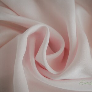 May include: A close-up of a soft, pale pink fabric with a smooth, silky texture. The fabric is draped and folded, creating a delicate and elegant look. The fabric is labeled "Emsilk".
