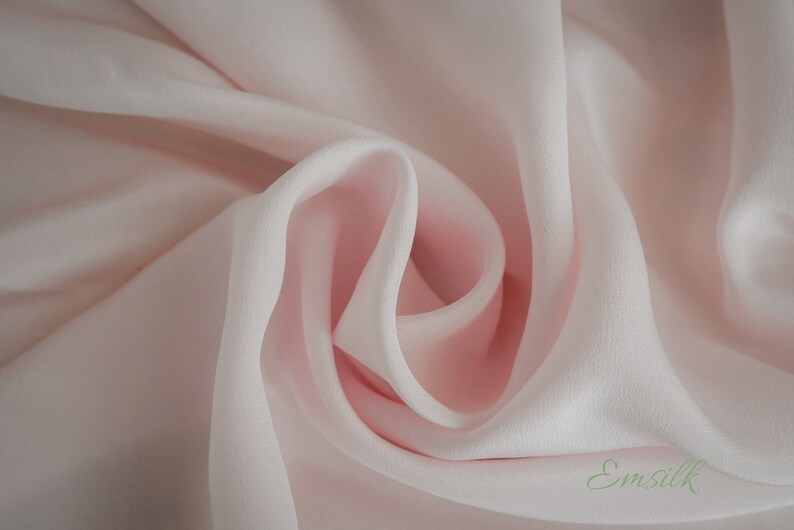 May include: A close-up of a soft, pale pink fabric with a smooth, silky texture. The fabric is draped and folded, creating a delicate and elegant look. The fabric is labeled "Emsilk".