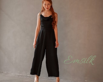 May include: A black wide-leg jumpsuit with a cowl neck and thin straps. The model is wearing the jumpsuit with gold heels and has long red hair.