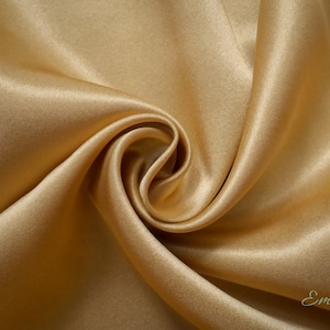 Op de afbeelding: Een close-up van een gladde, gouden satijnen stof. De stof is gedrapeerd en gevouwen, waardoor zachte, vloeiende lijnen ontstaan. De stof is luxueus en elegant.