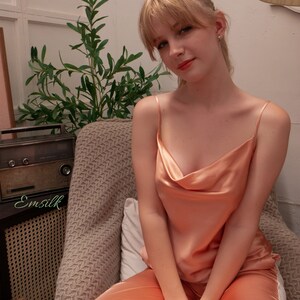 May include: A woman wearing a peach-coloured satin pyjama set with a cowl neck top. She is sitting in a chair with a plant and a vintage radio in the background.