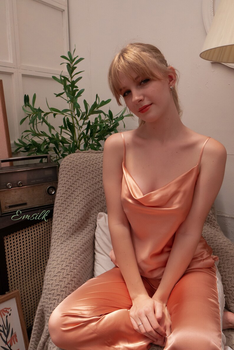 May include: A woman wearing a peach-coloured satin pyjama set with a cowl neck top. She is sitting in a chair with a plant and a vintage radio in the background.