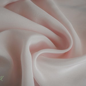 May include: A close-up of a soft, pale pink fabric with a smooth, silky texture. The fabric is draped and folded, creating a flowing, elegant look.