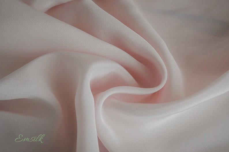 May include: A close-up of a soft, pale pink fabric with a smooth, silky texture. The fabric is draped and folded, creating a flowing, elegant look.
