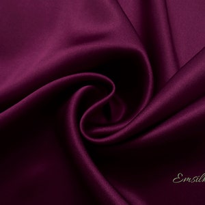 May include: Close-up of a deep purple satin fabric, showing its smooth texture and soft drape. The fabric is folded and arranged in a way that highlights its luxurious feel. The word "Emsilk" is visible in the bottom right corner.