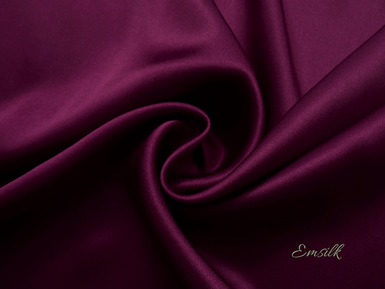 May include: Close-up of a deep purple satin fabric, showing its smooth texture and soft drape. The fabric is folded and arranged in a way that highlights its luxurious feel. The word "Emsilk" is visible in the bottom right corner.
