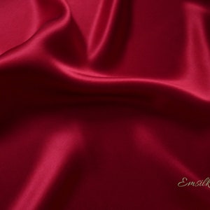May include: A close-up of a luxurious, flowing fabric in a deep burgundy color. The material has a silky sheen, with soft folds and highlights that create a sense of depth and movement. The word "Emsilk" is written in a cursive font in the bottom right corner.