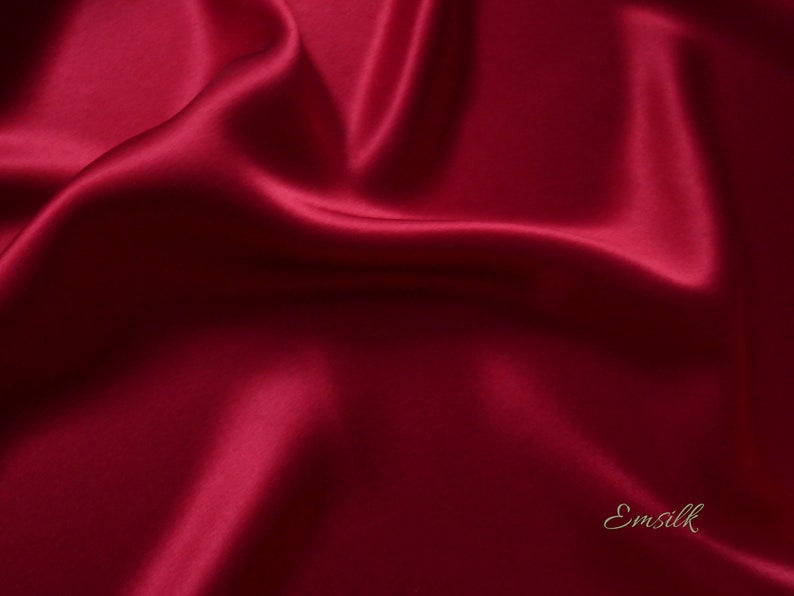 May include: A close-up of a luxurious, flowing fabric in a deep burgundy color. The material has a silky sheen, with soft folds and highlights that create a sense of depth and movement. The word "Emsilk" is written in a cursive font in the bottom right corner.