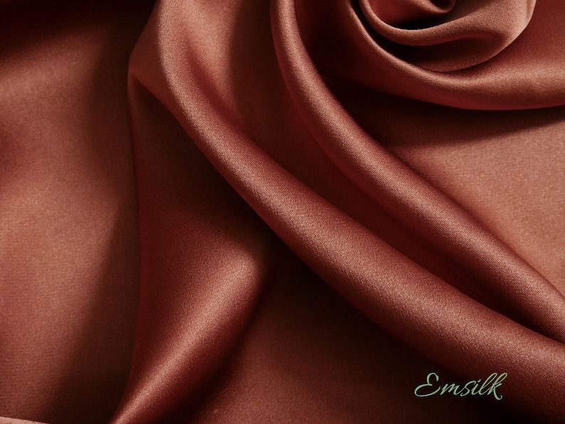 May include: Close-up of a luxurious, flowing fabric in a rich, warm terracotta color. The material has a subtle sheen, with soft folds and shadows that create depth. The fabric appears to be silk, with the word "Emsilk" in a cursive font.