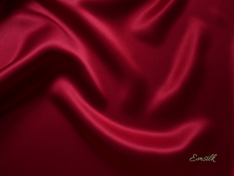 May include: A close-up of a deep red satin fabric with flowing folds and highlights. The fabric has a smooth, lustrous appearance, with shadows adding depth. The word "Emsilk" is in the bottom right corner.