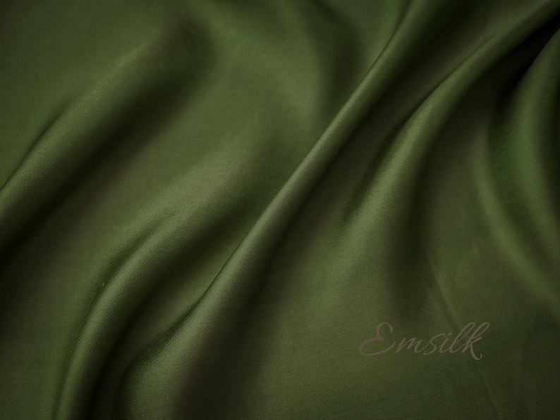 May include: A close-up of a smooth, dark green fabric with a subtle sheen. The fabric is draped in soft folds, creating a luxurious and elegant look. The word "Emsilk" is printed in a brown font on the fabric.