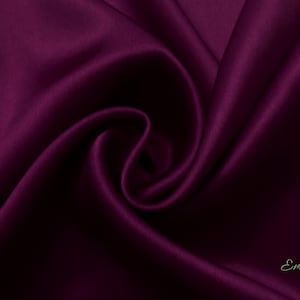 May include: A close-up of a deep purple satin fabric, showing the smooth texture and soft folds. The fabric is draped and folded, creating a luxurious and elegant look. The fabric is labeled "Emsilk" in the bottom right corner.