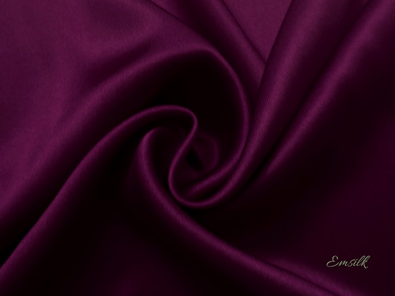 May include: A close-up of a deep purple satin fabric, showing the smooth texture and soft folds. The fabric is draped and folded, creating a luxurious and elegant look. The fabric is labeled "Emsilk" in the bottom right corner.