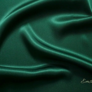 May include: A smooth, deep green fabric with a silky texture. The fabric is draped and folded, creating soft curves and folds. The word "Emsilk" is printed in a light green color at the bottom right corner of the image.