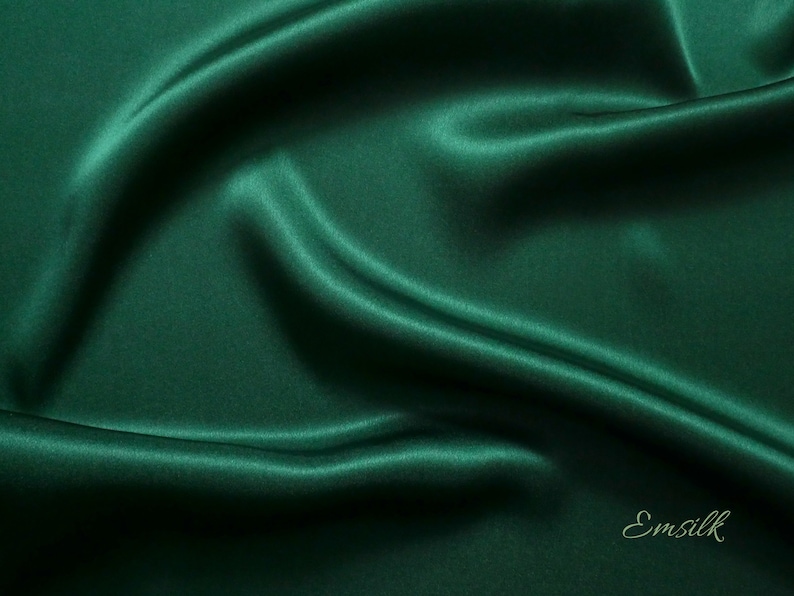 May include: A smooth, deep green fabric with a silky texture. The fabric is draped and folded, creating soft curves and folds. The word "Emsilk" is printed in a light green color at the bottom right corner of the image.