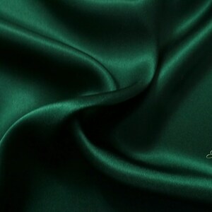 May include: A close-up of a deep green satin fabric, showing the smooth, luxurious texture and subtle sheen. The fabric is draped and folded, creating soft curves and folds.