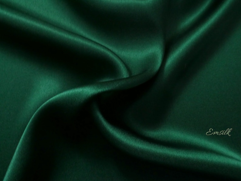 May include: A close-up of a deep green satin fabric, showing the smooth, luxurious texture and subtle sheen. The fabric is draped and folded, creating soft curves and folds.