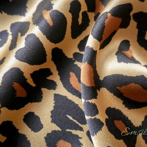 May include: Close-up of a beige silk fabric with a black and brown leopard print. The fabric is draped and has a soft, luxurious feel.
