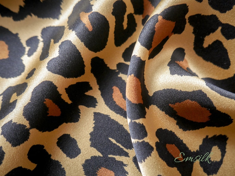May include: Close-up of a beige silk fabric with a black and brown leopard print. The fabric is draped and has a soft, luxurious feel.