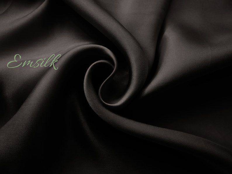 May include: Close-up of a smooth, black fabric with a subtle sheen. The fabric is draped in soft folds, creating a luxurious and elegant look. The text "Emsilk" is visible in the upper left corner.