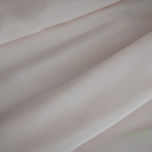 May include: A close-up of a pale pink fabric with a smooth, slightly textured surface. The fabric is folded in a way that creates soft, flowing lines.
