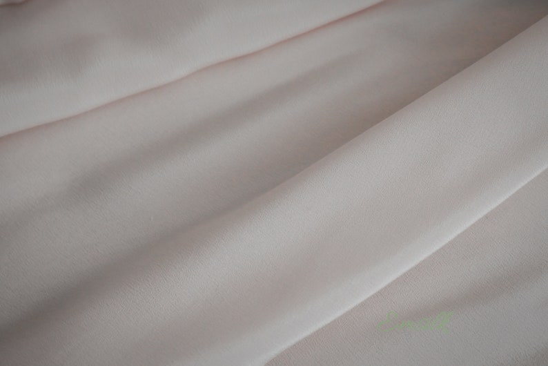 May include: A close-up of a pale pink fabric with a smooth, slightly textured surface. The fabric is folded in a way that creates soft, flowing lines.