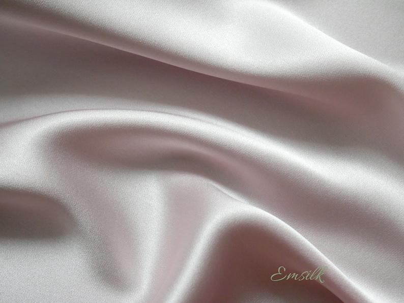 May include: A close-up of a smooth, pale pink satin fabric with a subtle sheen. The fabric is draped and folded, creating soft curves and folds.
