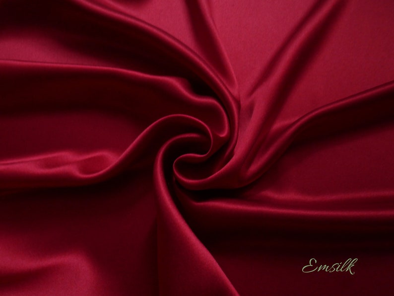 May include: Close-up of a deep red satin fabric with elegant folds and a swirling pattern. The fabric has a lustrous sheen, reflecting light and creating highlights and shadows. The word "Emsilk" is in the bottom right corner.