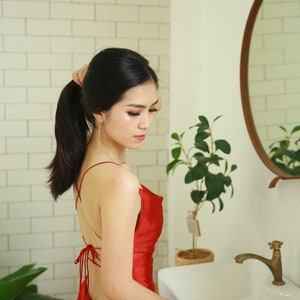 May include: A woman with long black hair tied back in a ponytail is wearing a red satin dress with a low back and a criss-cross strap design. She is standing in a bathroom with a white sink and a mirror.