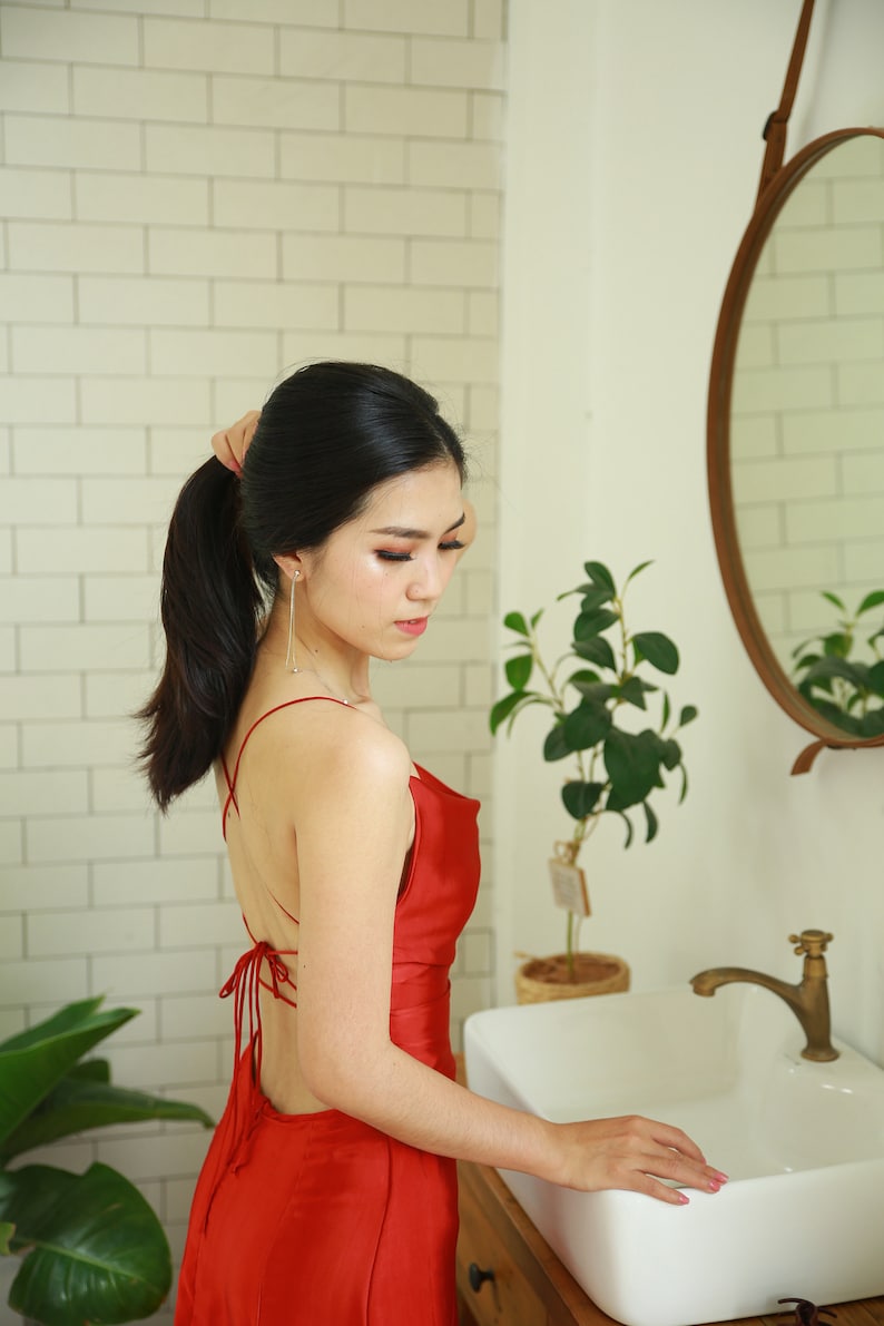 May include: A woman with long black hair tied back in a ponytail is wearing a red satin dress with a low back and a criss-cross strap design. She is standing in a bathroom with a white sink and a mirror.