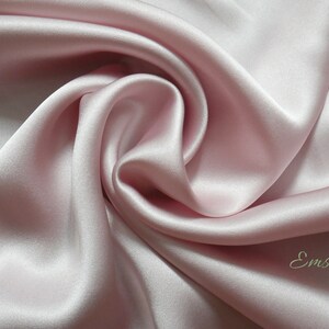 May include: A close-up of a soft, smooth, pale pink silk fabric. The fabric is draped and folded, creating a luxurious and elegant look. The fabric is labeled "Emsilk".