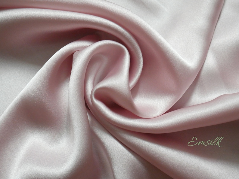 May include: A close-up of a soft, smooth, pale pink silk fabric. The fabric is draped and folded, creating a luxurious and elegant look. The fabric is labeled "Emsilk".
