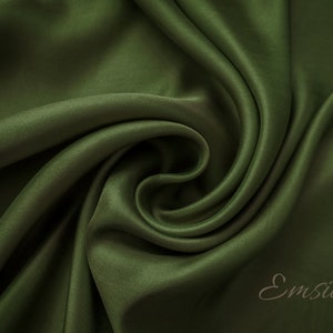May include: Close-up of a deep green satin fabric, showing its smooth texture and soft drape. The fabric is folded and twisted, creating a swirling pattern. The word "Emsilk" is visible in the lower right corner.