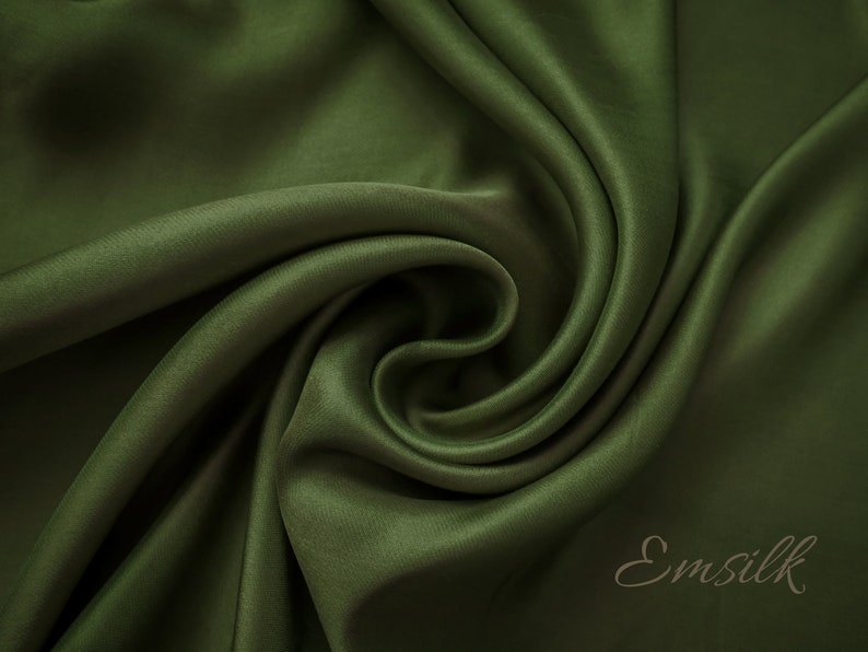 May include: Close-up of a deep green satin fabric, showing its smooth texture and soft drape. The fabric is folded and twisted, creating a swirling pattern. The word "Emsilk" is visible in the lower right corner.