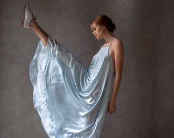 May include: A woman wearing a light blue maxi dress with a halter neckline and a flowing skirt. She is standing on one leg with her other leg raised in the air. She is wearing white sneakers. The background is a gray wall.