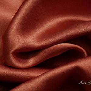 May include: Close-up of a luxurious, flowing fabric in a rich, warm reddish-brown hue. The material has a smooth, lustrous surface with soft folds and shadows, suggesting a high-quality silk or satin textile. The word "Emsilk" is in the bottom right corner.