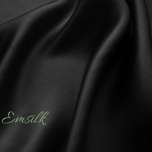 May include: A close-up of a smooth, black fabric with a subtle sheen. The fabric is draped in soft folds, creating a luxurious and elegant look. The word "Emsilk" is written in a cursive font on the fabric.