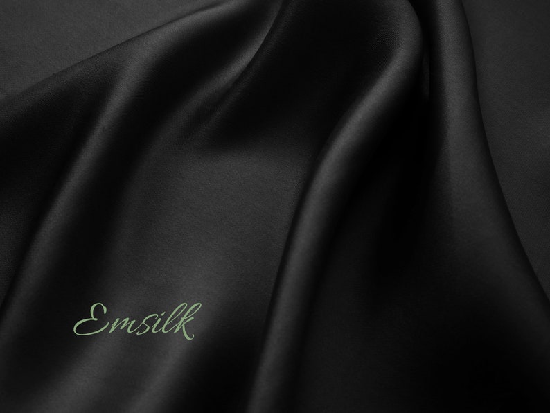 May include: A close-up of a smooth, black fabric with a subtle sheen. The fabric is draped in soft folds, creating a luxurious and elegant look. The word "Emsilk" is written in a cursive font on the fabric.
