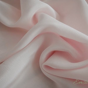 May include: A close-up of a soft, pale pink fabric with a smooth, silky texture. The fabric is draped and folded, creating a luxurious and elegant look.
