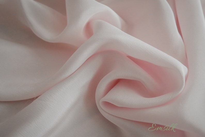 May include: A close-up of a soft, pale pink fabric with a smooth, silky texture. The fabric is draped and folded, creating a luxurious and elegant look.