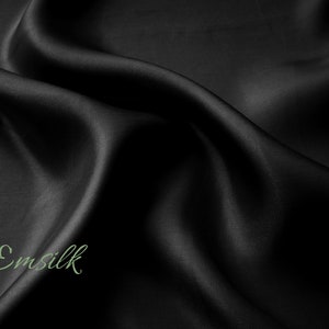 May include: Black satin fabric with a smooth, silky texture. The fabric is draped and folded, creating soft curves and folds. The word "Emsilk" is written in green script on the fabric.