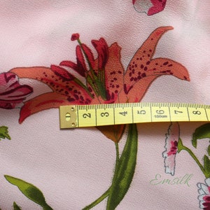 May include: Pink fabric with a floral print featuring a large orange lily with red markings and smaller pink flowers. A yellow measuring tape is placed across the lily, showing measurements in inches.